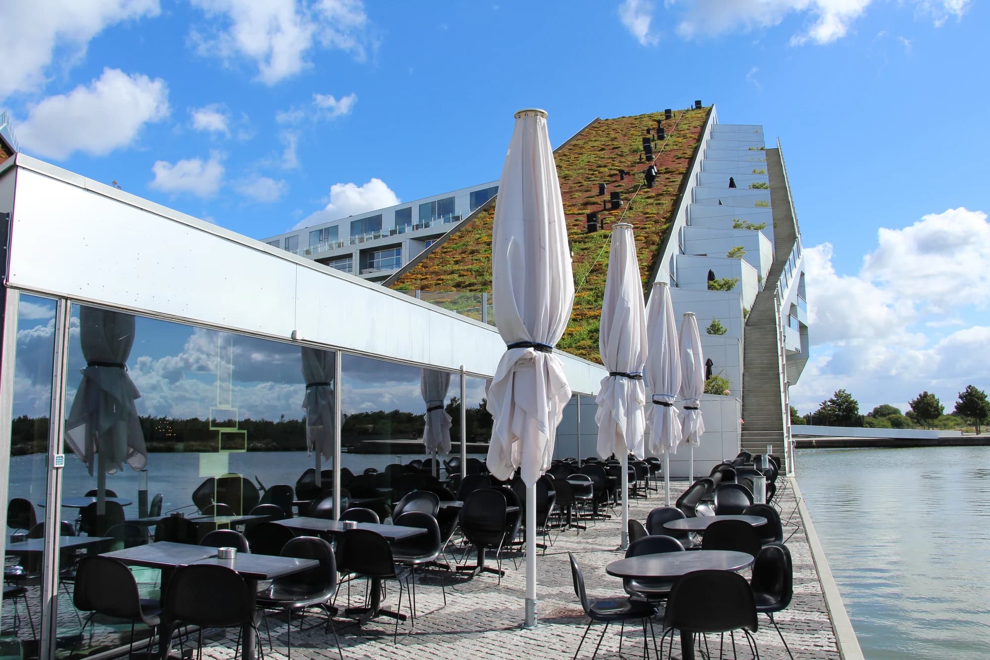 Outdoor restaurant tables beside the water at 8 House, with the sloping residential building and balconies behind.